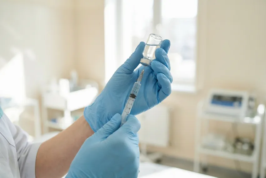 Roke strokovnjaka v zaščitnih rokavicah držijo brizgo in vialo v svetli ambulanti.